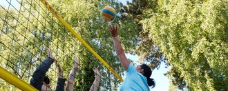 Action: eine Gruppe Frauen und Männer spielen Volleyball - eine Frau "lupft" gerade den Ball über den "Block"