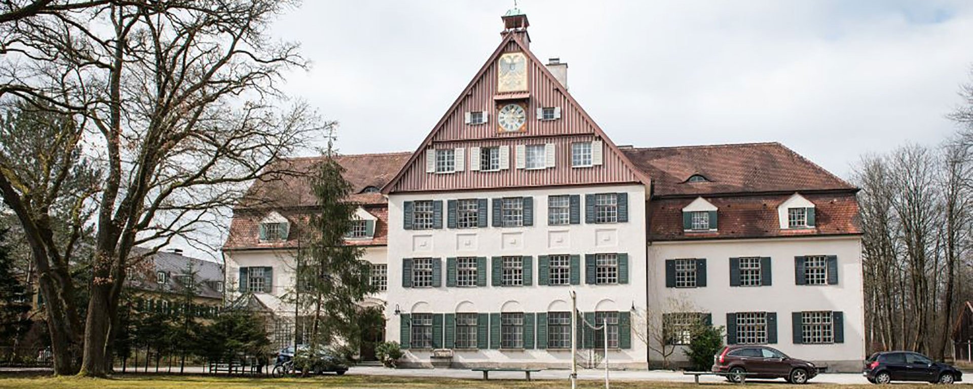 Aussenaufnahme: Gebäude mit großer Uhr an der Stirnseite - viele grüne Fensteläden, Bäume und ein Volleyballnetz im Vordergrund