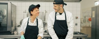Zwei Menschen stehen in der Küche, schneiden Gemüse und lachen sich an.
