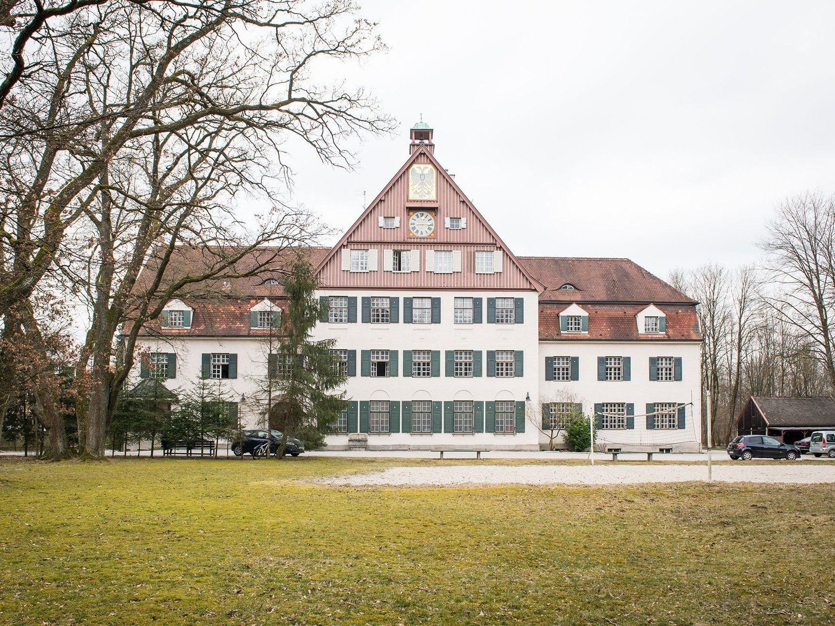 Aussenaufnahme: Gebäude mit großer Uhr an der Stirnseite - viele grüne Fensteläden, Bäume und ein Volleyballnetz im Vordergrund