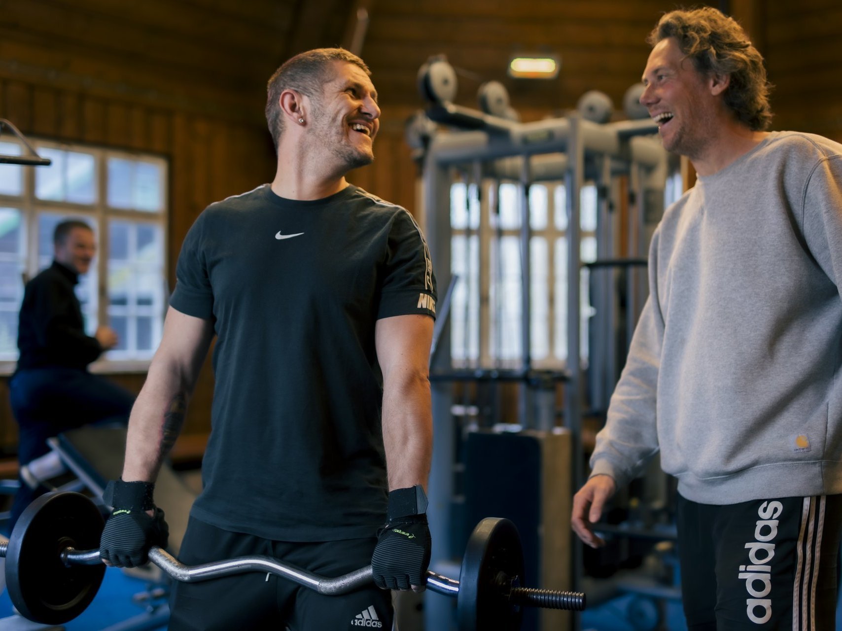 Ein Mann hält eine Hantel in den Händen und lacht seinen Trainer an, der schräg hinter ihm steht.