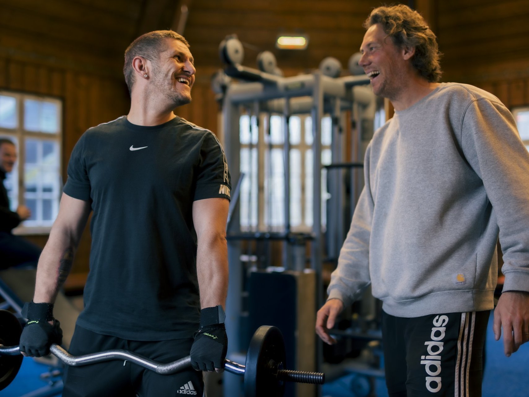 Ein Mann hält eine Hantel in den Händen und lacht seinen Trainer an, der schräg hinter ihm steht.