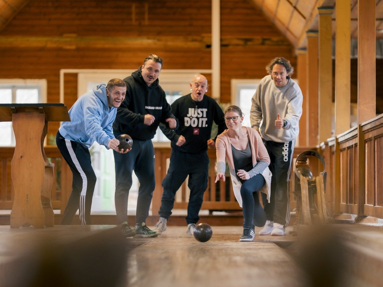 Fünf Menschen stehen am Kopf einer Bowlingbahn und feuern eine Frau an, die eine Bowlingkugel wirft.
