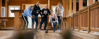 Fünf Menschen stehen am Kopf einer Bowlingbahn und feuern eine Frau an, die eine Bowlingkugel wirft.